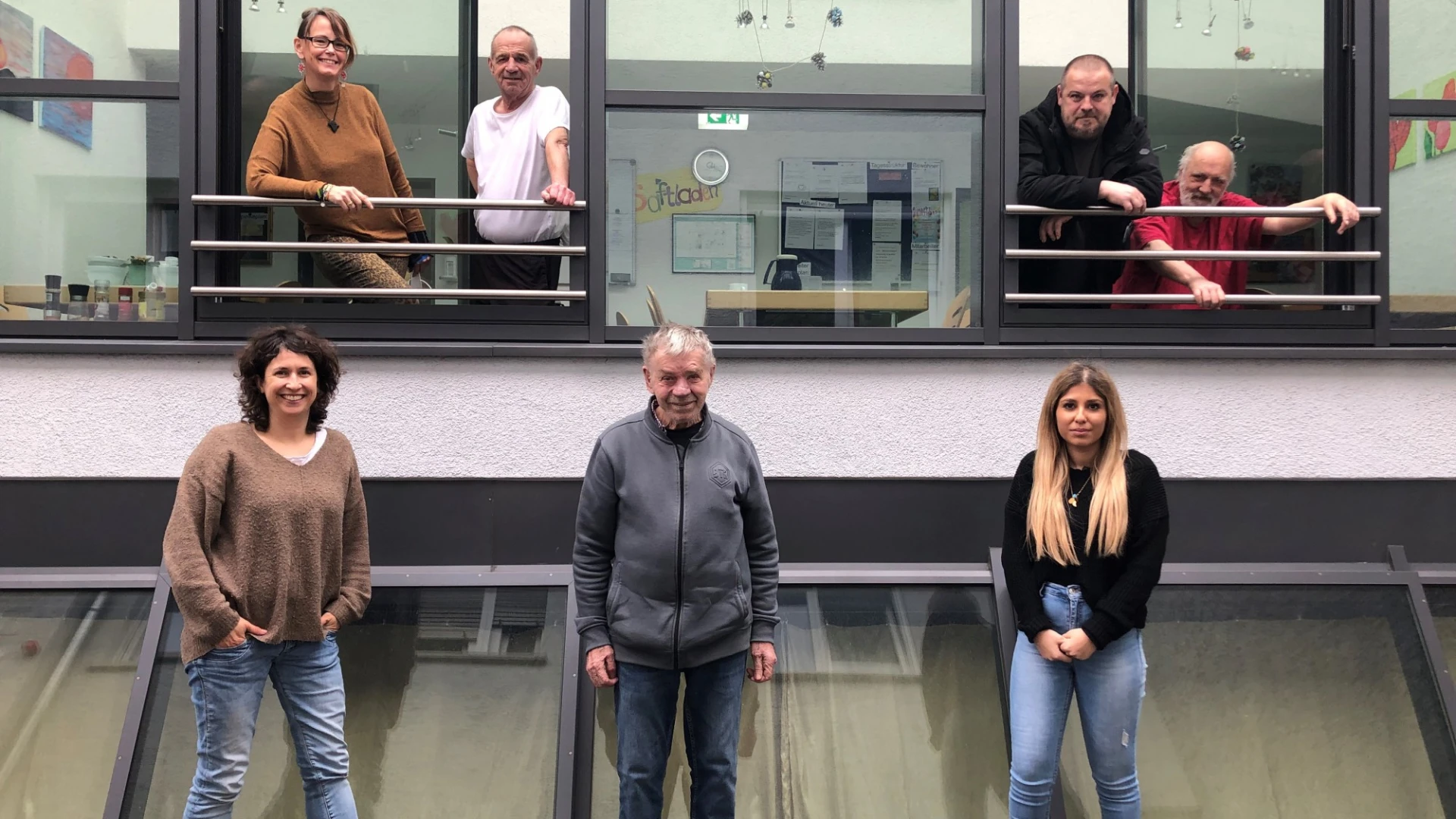 Drei Personen sind vor einem Geb&auml;ude zu sehen, vier stehen in zwei ge&ouml;ffneten Fenstern im oberen Stockwerk.