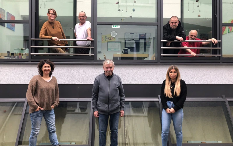 Drei Personen sind vor einem Geb&auml;ude zu sehen, vier stehen in zwei ge&ouml;ffneten Fenstern im oberen Stockwerk.
