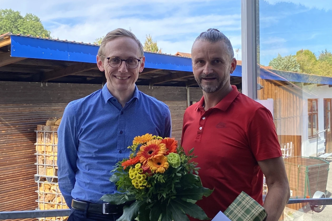 Zwei M&auml;nner l&auml;cheln in die Kamera. Einem wurde ein Geschenk und ein Blumenstrau&szlig; &uuml;berreicht.