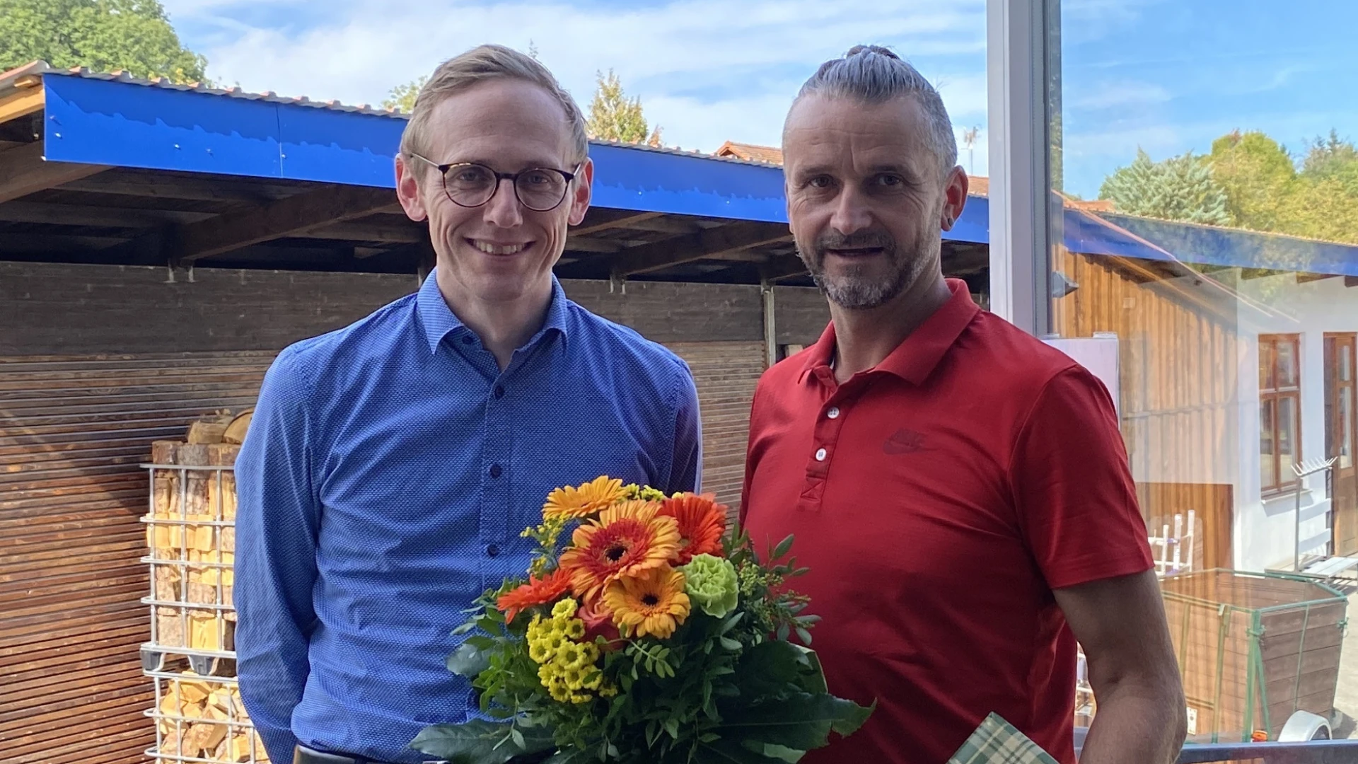 Zwei M&auml;nner l&auml;cheln in die Kamera. Einem wurde ein Geschenk und ein Blumenstrau&szlig; &uuml;berreicht.
