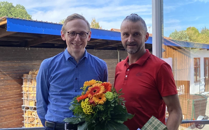 Zwei M&auml;nner l&auml;cheln in die Kamera. Einem wurde ein Geschenk und ein Blumenstrau&szlig; &uuml;berreicht.