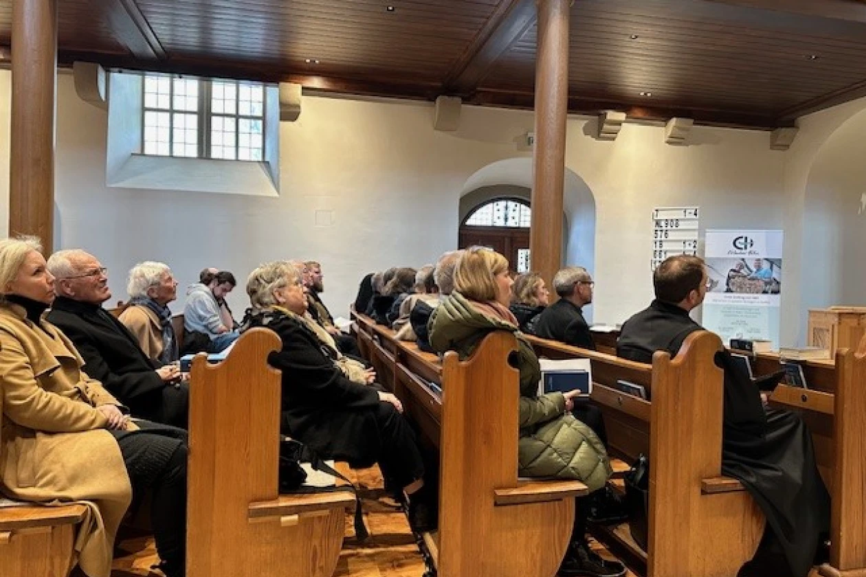 Mehrere Personen sitzen auf Holzb&auml;nken in einem Kirchenraum mit Fenstern. Neben der Eingangst&uuml;r ist ein Roll-up der Erlacher H&ouml;he zu sehen. Die Personen tragen warme Winterkleidung, und blicken nach vorne.
