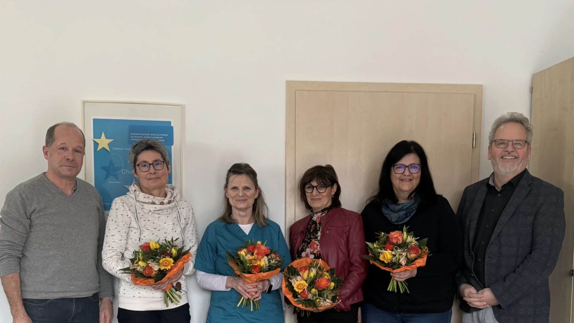 Vier Frauen mit Blumenstrau&szlig; stehen zwischen zwei M&auml;nnern. Alle blicken in die Kamera. Im Vordergrund steht ein Tisch mit einem weiteren Blumenstrau&szlig;.