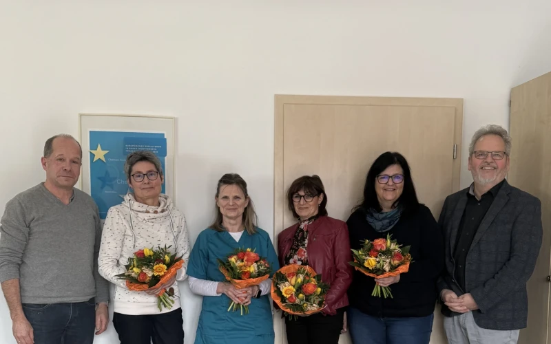 Vier Frauen mit Blumenstrau&szlig; stehen zwischen zwei M&auml;nnern. Alle blicken in die Kamera. Im Vordergrund steht ein Tisch mit einem weiteren Blumenstrau&szlig;.