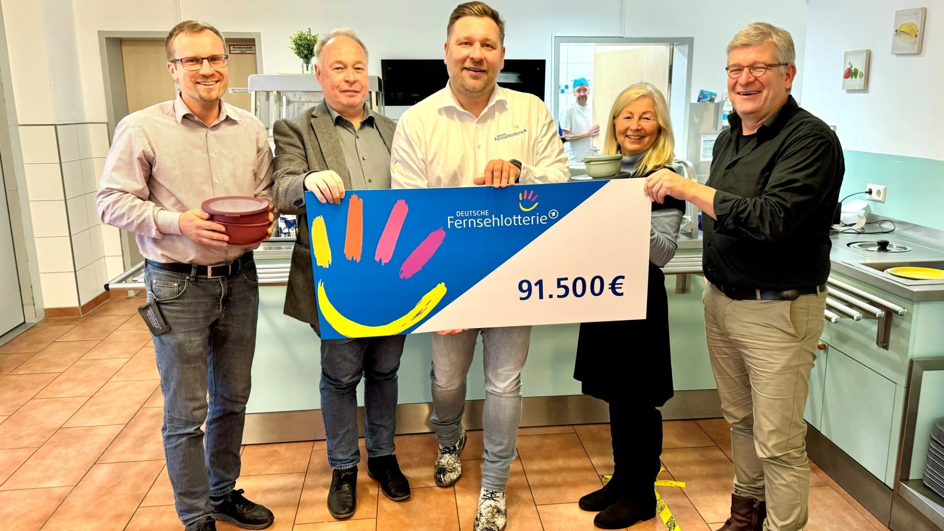 Vier M&auml;nner und eine Frau l&auml;cheln freudig in die Kamera. Ein F&ouml;rdergutschein der Fernsehlotterie wird &uuml;bergeben.