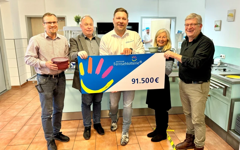 Vier M&auml;nner und eine Frau l&auml;cheln freudig in die Kamera. Ein F&ouml;rdergutschein der Fernsehlotterie wird &uuml;bergeben.