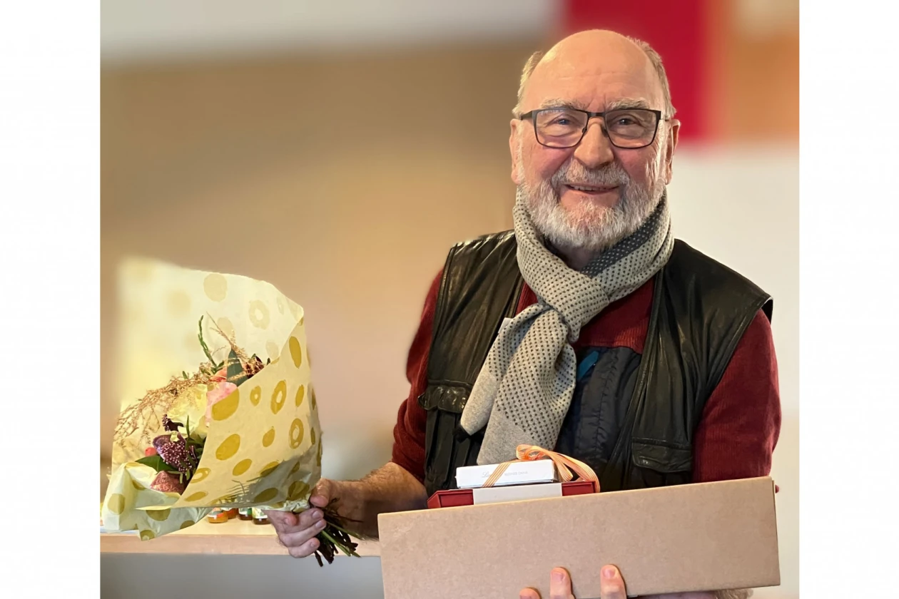 Ein Mann mit Brille und grauem Bart tr&auml;gt eine schwarze Weste &uuml;ber einem roten Hemd und einen grauen Schal. Er h&auml;lt eine Schachtel und einen Blumenstrau&szlig; in den H&auml;nden und l&auml;chelt in die Kamera