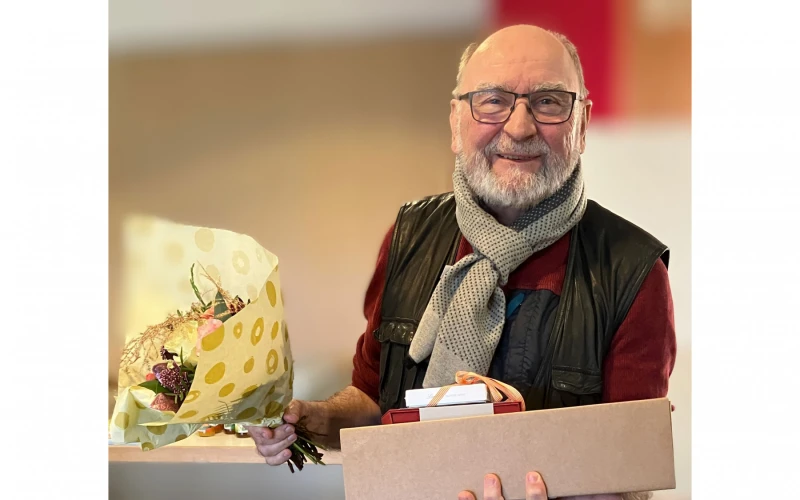 Ein Mann mit Brille und grauem Bart tr&auml;gt eine schwarze Weste &uuml;ber einem roten Hemd und einen grauen Schal. Er h&auml;lt eine Schachtel und einen Blumenstrau&szlig; in den H&auml;nden und l&auml;chelt in die Kamera