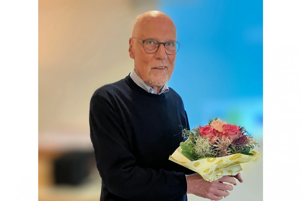 Ein Mann mit Brille tr&auml;gt einen dunklen Pullover und h&auml;lt einen Blumenstrau&szlig; mit roten und rosa Bl&uuml;ten in den H&auml;nden.