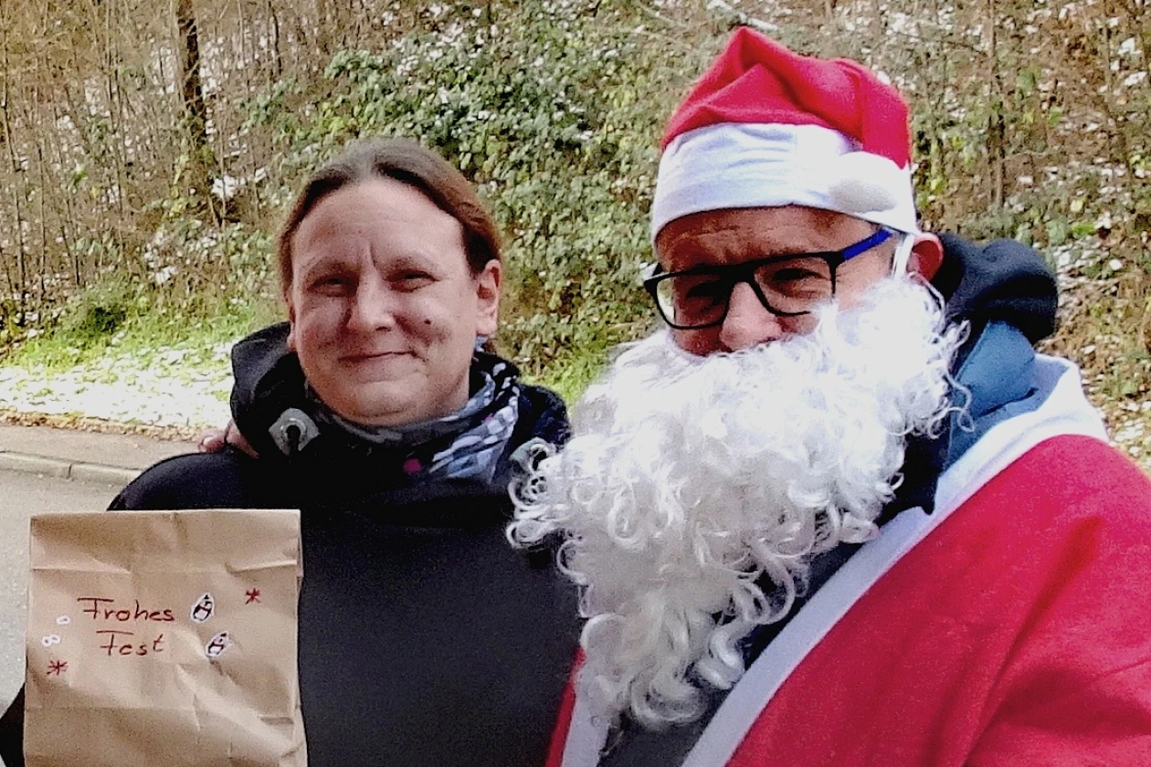 Zwei Menschen im Freien: Eine Frau in schwarzer Winterkleidung h&auml;lt einen Papierbeutel mit der Aufschrift "Frohes Fest", w&auml;hrend die andere Person als Weihnachtsmann mit roter Jacke, wei&szlig;em Bart und M&uuml;tze verkleidet ist.