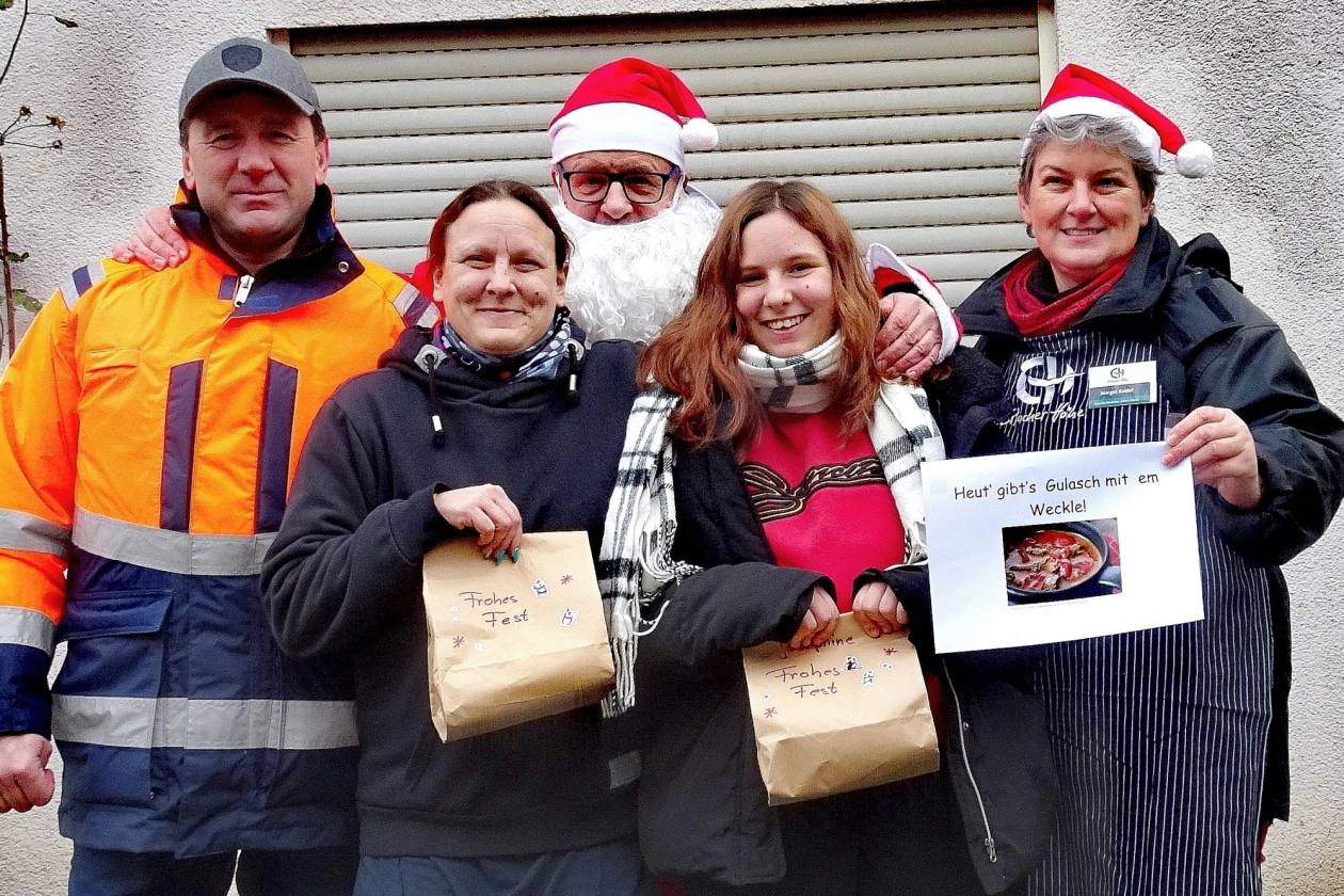 F&uuml;nf Personen in Winterkleidung, darunter ein Weihnachtsmann, stehen zusammen und halten Geschenkpakete. Sie tragen weihnachtliche M&uuml;tzen und scheinen an einer Weihnachtsaktion teilzunehmen, vor einer Rolltor-Kulisse.