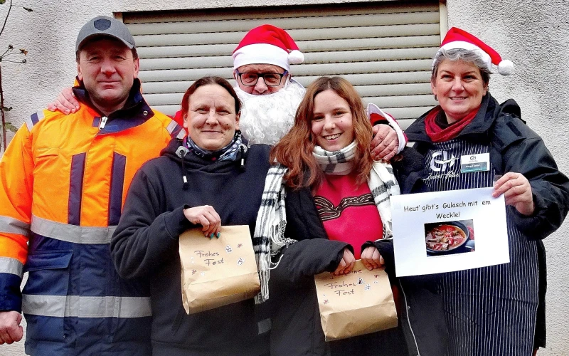F&uuml;nf Personen in Winterkleidung, darunter ein Weihnachtsmann, stehen zusammen und halten Geschenkpakete. Sie tragen weihnachtliche M&uuml;tzen und scheinen an einer Weihnachtsaktion teilzunehmen, vor einer Rolltor-Kulisse.