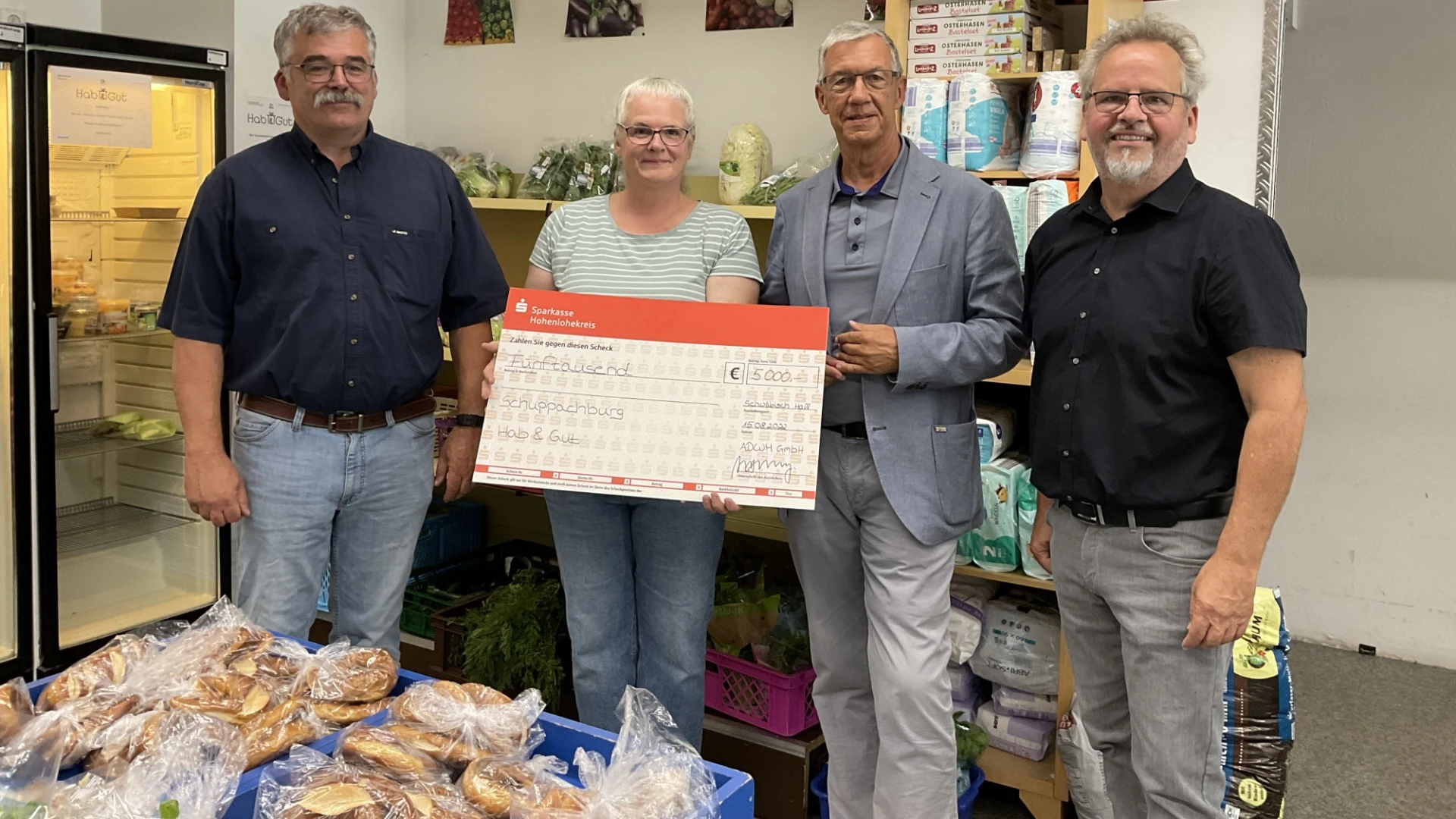 Drei M&auml;nner und eine Frau blicken in die Kamera. Im Vordergrund sind eingepackte Backwaren zu sehen, im Hintergrund ein K&uuml;hlschrank und Lebensmittelregale. Die Frau h&auml;lt einen Spendenscheck in der Hand.