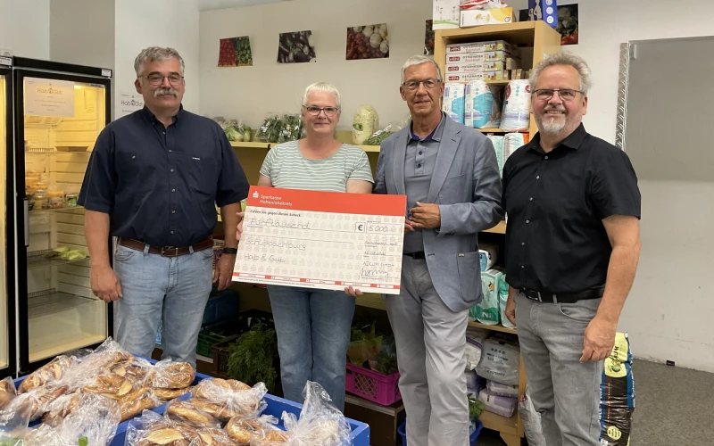 Drei M&auml;nner und eine Frau blicken in die Kamera. Im Vordergrund sind eingepackte Backwaren zu sehen, im Hintergrund ein K&uuml;hlschrank und Lebensmittelregale. Die Frau h&auml;lt einen Spendenscheck in der Hand.