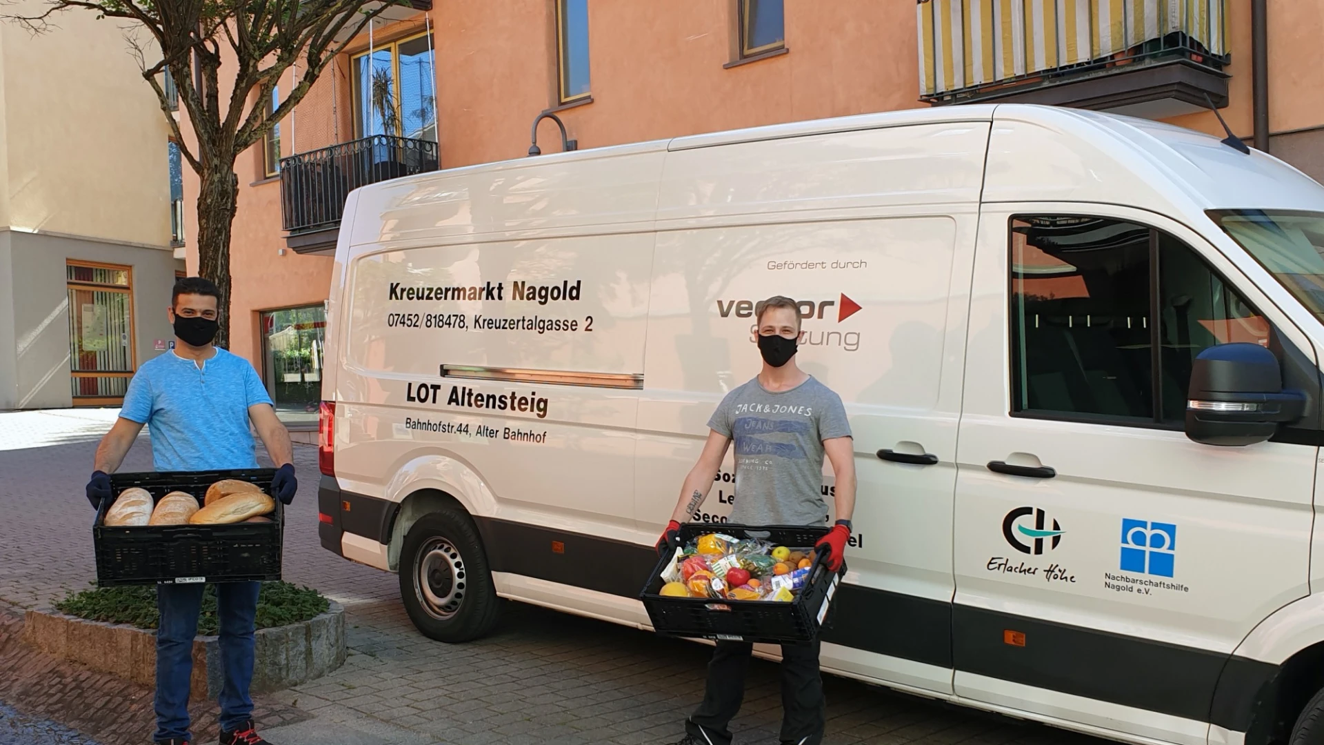 Zwei M&auml;nner stehen vor einem wei&szlig;en Transporter mit der Aufschrift &sbquo;Kreuzermarkt Nagold&lsquo; sowie weiteren Beschriftungen. Beide tragen schwarze Kisten mit Lebensmitteln und sind mit schwarzen Mund-Nasenschutz und Handschuhen ausgestattet.