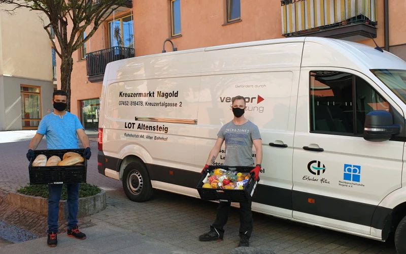 Zwei M&auml;nner stehen vor einem wei&szlig;en Transporter mit der Aufschrift &sbquo;Kreuzermarkt Nagold&lsquo; sowie weiteren Beschriftungen. Beide tragen schwarze Kisten mit Lebensmitteln und sind mit schwarzen Mund-Nasenschutz und Handschuhen ausgestattet.