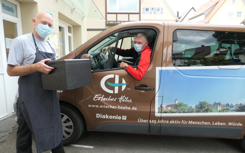 Ein Mann sitzt in einem braunen Lieferwagen, der mit dem Logo der Erlacher H&ouml;he beklebt ist. Neben ihm steht ein Mann mit Sch&uuml;rze und einer schwarzen Lieferbox. Beide tragen Mund-Nasenschutz.