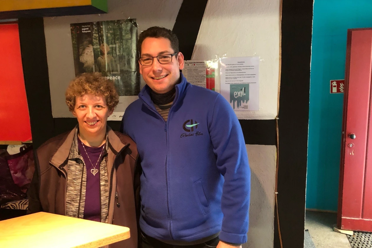 Zwei l&auml;chelnde Personen stehen in einem Raum vor einer Wand. Die Frau mit lockigen Haaren tr&auml;gt eine braune Jacke, der j&uuml;ngere Mann neben ihr tr&auml;gt eine blaue Fleecejacke und eine Brille. Im Hintergrund sind Zettel an der Wand zu sehen, sowie eine rote, ge&ouml;ffnete T&uuml;r.