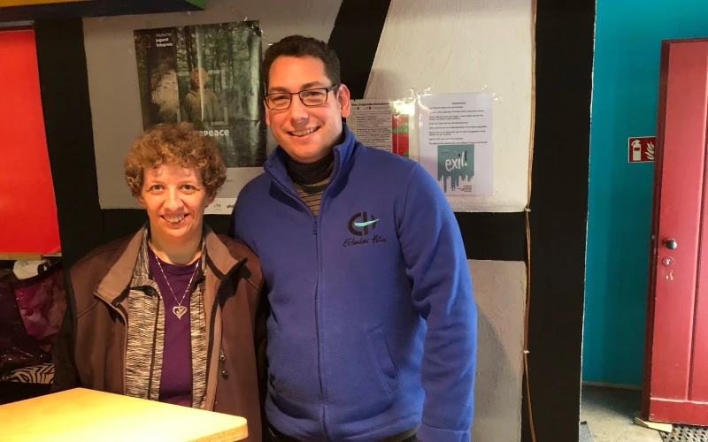 Zwei l&auml;chelnde Personen stehen in einem Raum vor einer Wand. Die Frau mit lockigen Haaren tr&auml;gt eine braune Jacke, der j&uuml;ngere Mann neben ihr tr&auml;gt eine blaue Fleecejacke und eine Brille. Im Hintergrund sind Zettel an der Wand zu sehen, sowie eine rote, ge&ouml;ffnete T&uuml;r.