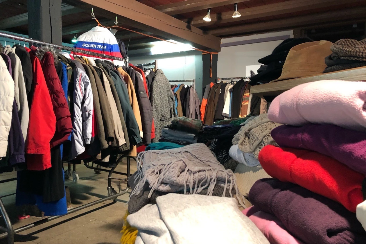 Ein Lagerraum mit gespendeten Winterkleidungsst&uuml;cken, darunter M&auml;ntel, Jacken und Pullover auf Kleiderstangen und Stapeln