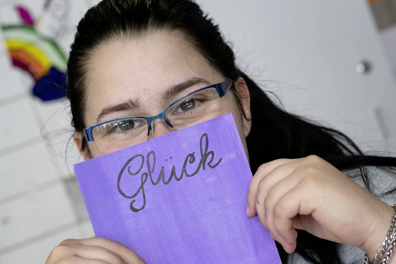 Eine junge Frau mit Brille und dunklen langen Haaren tr&auml;gt ein graues T-Shirt mit dem Aufdruck "Live the life you". Sie h&auml;lt eine lila Karte mit dem Wort "Gl&uuml;ck" vor ihr Gesicht. Sie sitzt an einem Tisch mit Dokumenten und l&auml;chelt in die Kamera.