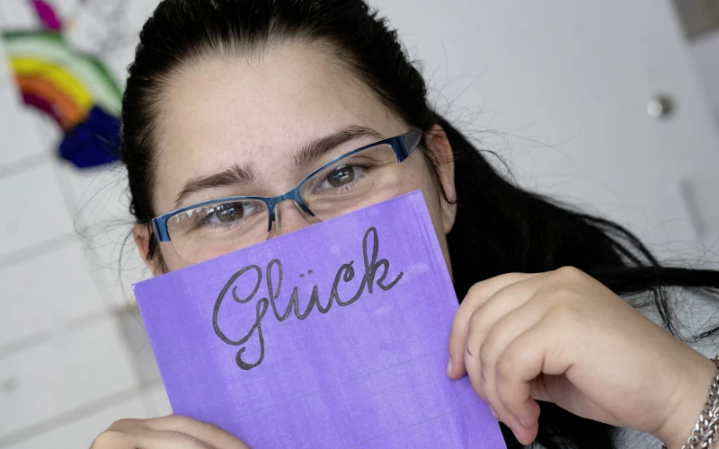 Eine junge Frau mit Brille und dunklen langen Haaren tr&auml;gt ein graues T-Shirt mit dem Aufdruck "Live the life you". Sie h&auml;lt eine lila Karte mit dem Wort "Gl&uuml;ck" vor ihr Gesicht. Sie sitzt an einem Tisch mit Dokumenten und l&auml;chelt in die Kamera.