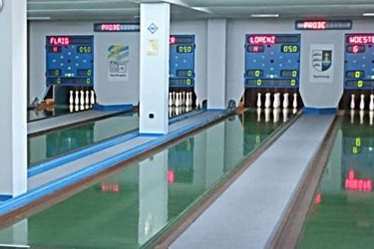 Vier Bowlingbahnen mit gr&uuml;nem Boden, ausgestattet mit elektronischen Anzeigetafeln an der Wand sind zu sehen. Wei&szlig;e Bowlingkegel stehen am Ende jeder Bahn bereit zum Spiel.