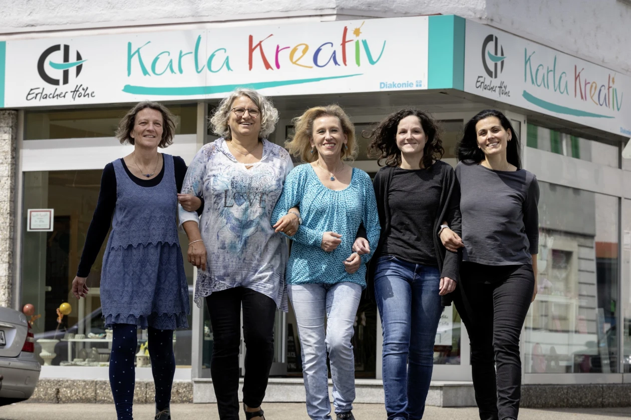 F&uuml;nf Frauen laufen freudig und eingehakt vor dem Gesch&auml;ft "Karla Kreativ" auf die Kamera zu. Sie tragen verschiedene Kleidungsst&uuml;cke in Blau-, Grau- und Schwarzt&ouml;nen. Der Hintergrund zeigt die Ladenfront mit dem bunten Schriftzug und dem Logo der Erlacher H&ouml;he.