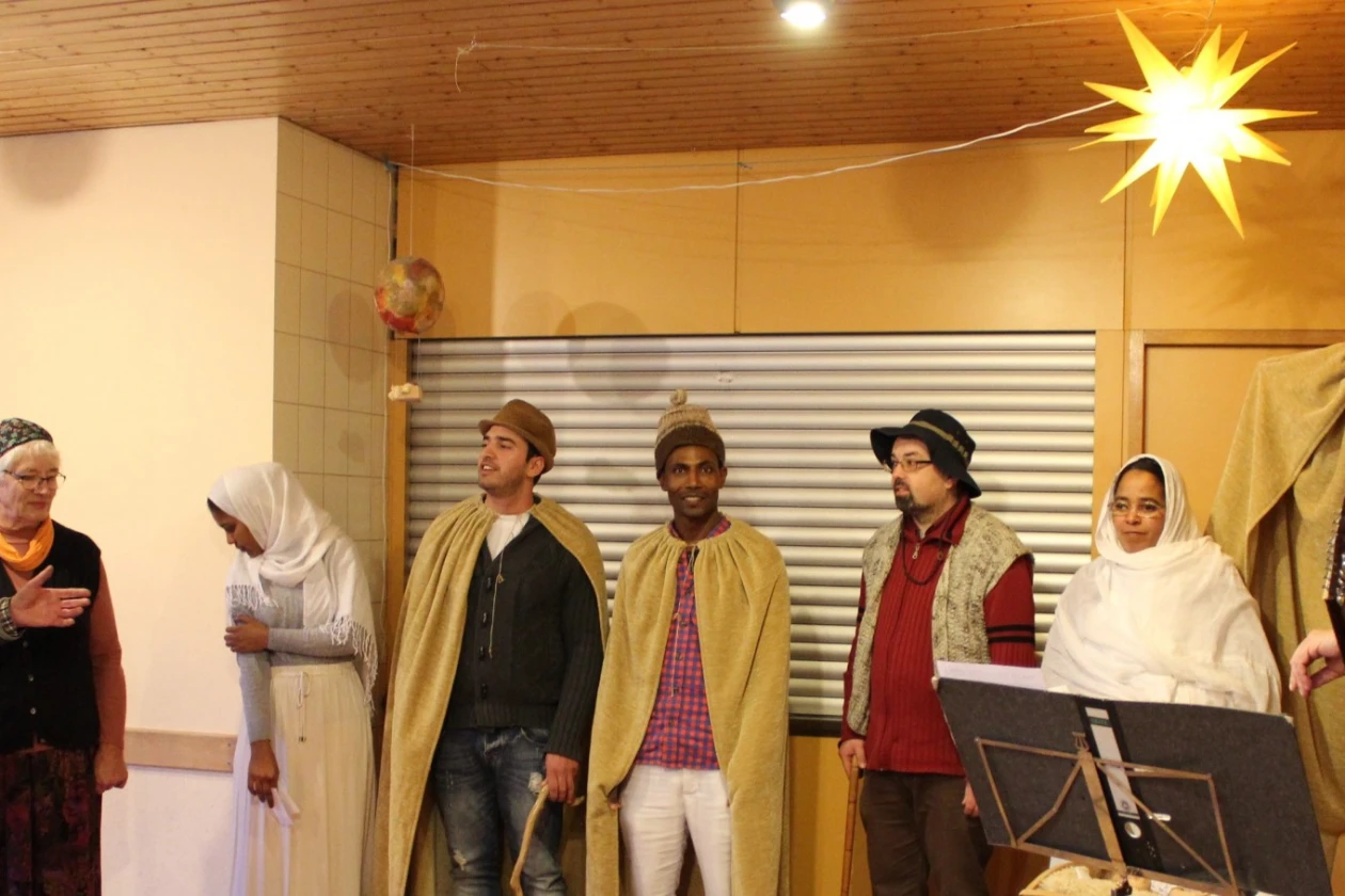 Eine Gruppe von acht Personen stellt eine weihnachtliche Szene in einem Raum unter einem hellen Stern. Der Mann auf der rechten Seite h&auml;lt eine Gitarre in der Hand.