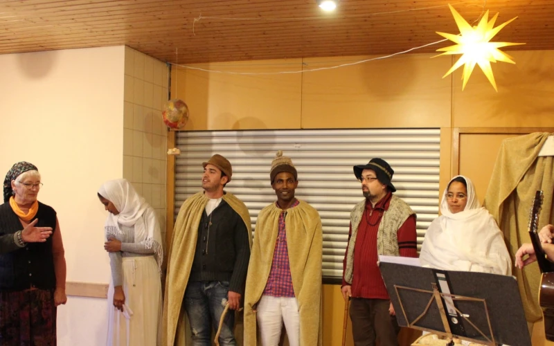 Eine Gruppe von acht Personen stellt eine weihnachtliche Szene in einem Raum unter einem hellen Stern. Der Mann auf der rechten Seite h&auml;lt eine Gitarre in der Hand.