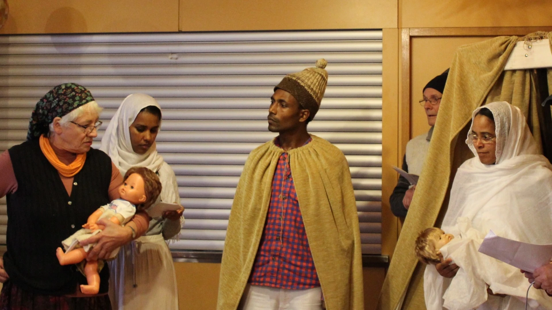 Weihnachtliche Szene mit mehreren Personen in traditioneller Kleidung, mit einer Krippe, einem hellen Stern und einer Frau, die ein Baby auf dem Arm h&auml;lt.