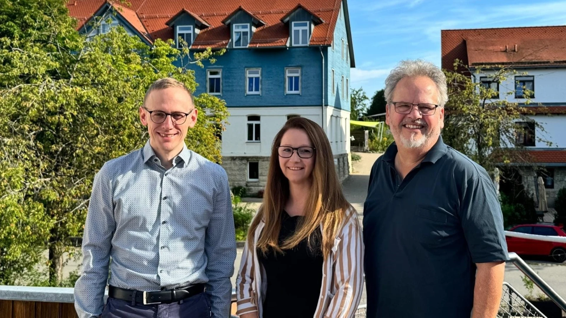Eine Frau und zwei M&auml;nner blicken l&auml;chelnd in die Kamera. Im Hintergrund sind weitere Geb&auml;ude und B&auml;ume zu sehen.