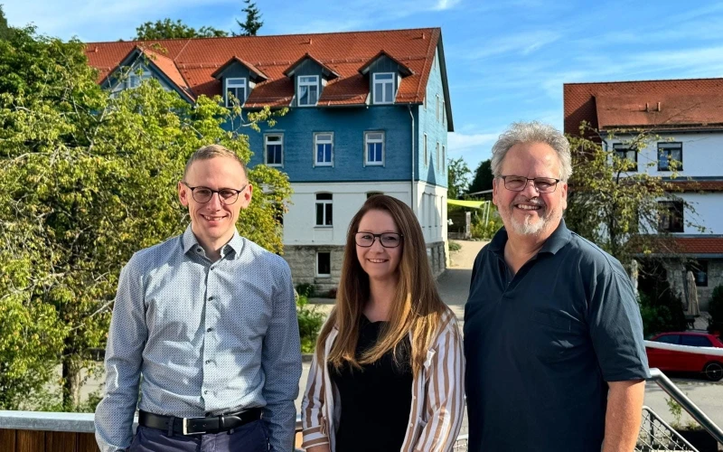 Eine Frau und zwei M&auml;nner blicken l&auml;chelnd in die Kamera. Im Hintergrund sind weitere Geb&auml;ude und B&auml;ume zu sehen.