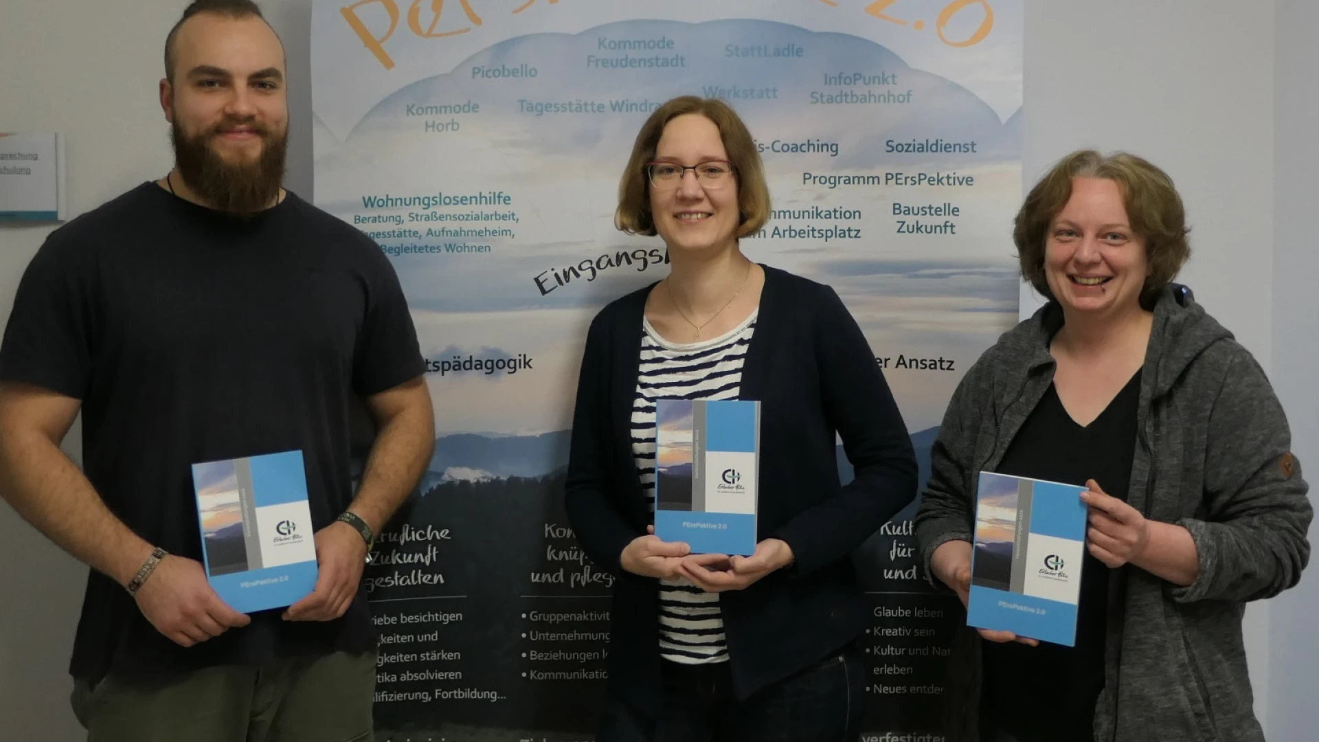 Drei Personen stehen vor einem Roll-up mit dem Titel "Perspektive 2.0", das zahlreiche Informationen zeigt. Jede der drei Personen h&auml;lt ein Buch mit dem Logo der Erlacher H&ouml;he in der Hand.