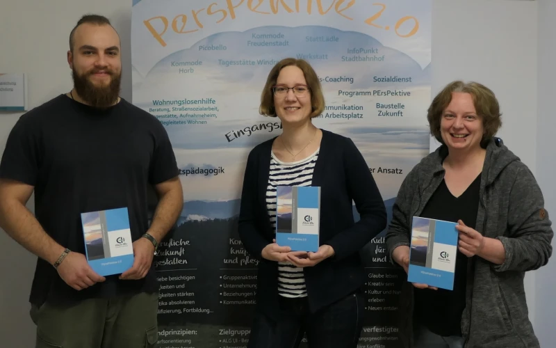 Drei Personen stehen vor einem Roll-up mit dem Titel "Perspektive 2.0", das zahlreiche Informationen zeigt. Jede der drei Personen h&auml;lt ein Buch mit dem Logo der Erlacher H&ouml;he in der Hand.