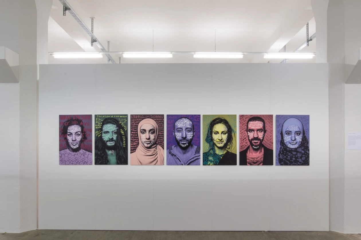 Sieben gro&szlig;formatige Portr&auml;ts in verschiedenen Farbt&ouml;nen h&auml;ngen an einer wei&szlig;en Wand eines Ausstellungsraums. Die gerahmten Bilder zeigen Gesichter mit unterschiedlichen Ausdr&uuml;cken und Hintergr&uuml;nden, die in Lila-, Gr&uuml;n-, Rosa- und Blaut&ouml;nen gehalten sind.