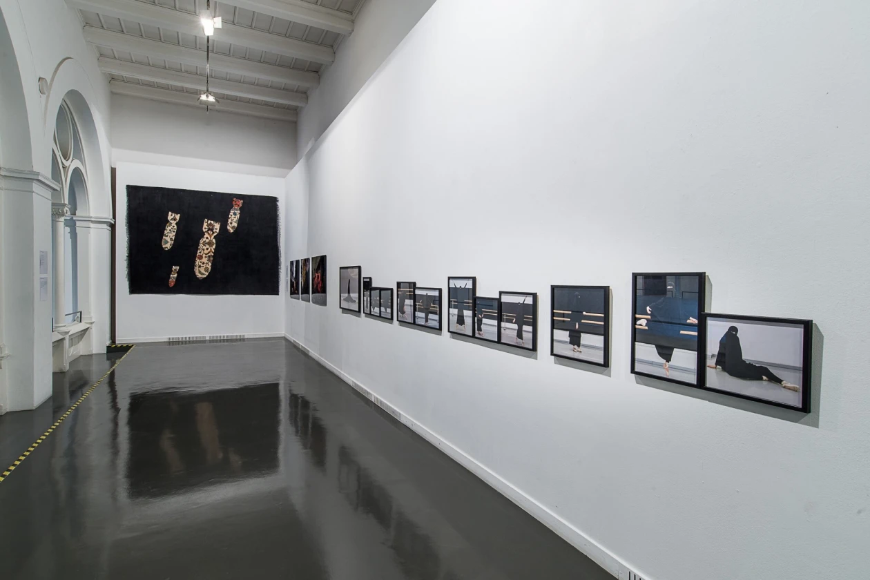 Ein Museumsraum mit wei&szlig;en W&auml;nden und schwarzem Boden. An der rechten Wand h&auml;ngen mehrere in schwarz gerahmte Fotografien in unterschiedlichen Gr&ouml;&szlig;en. Ein schwarzes Kunstwerk dominiert die linke Wand.