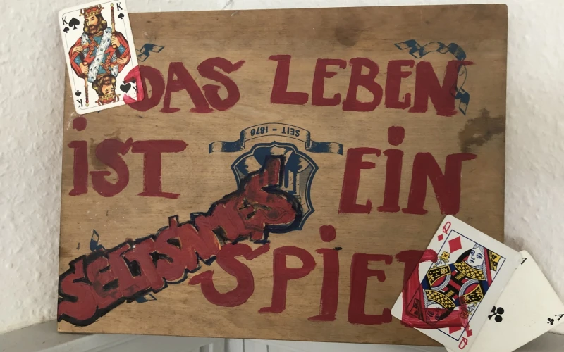 Das Kunstwerk zeigt auf einem Holzbrett den Text "Das Leben ist ein seltsames Spiel", aufgeklebt sind Spielkarten.