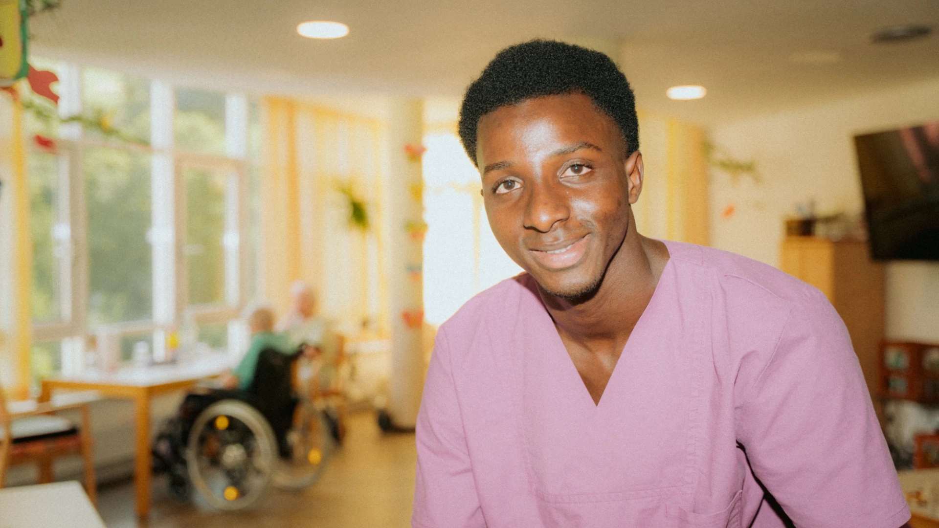 Ein l&auml;chelnder Mann in pinkem medizinischem Kittel steht in einer hellen Einrichtung mit Behandlungszimmern im Hintergrund