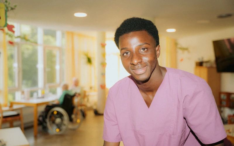 Ein l&auml;chelnder Mann in pinkem medizinischem Kittel steht in einer hellen Einrichtung mit Behandlungszimmern im Hintergrund