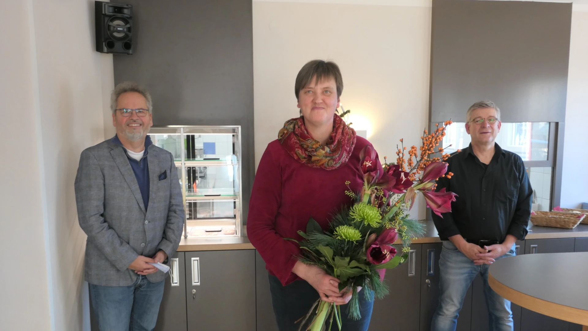 Drei Personen stehen in einem Bistro und blicken freundlich in die Kamera. Die Frau in der Mitte h&auml;lt einen Blumenstrau&szlig; in den H&auml;nden. Rechts im Vordergrund ist ein runder Stehtisch zu sehen.