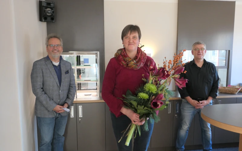 Drei Personen stehen in einem Bistro und blicken freundlich in die Kamera. Die Frau in der Mitte h&auml;lt einen Blumenstrau&szlig; in den H&auml;nden. Rechts im Vordergrund ist ein runder Stehtisch zu sehen.