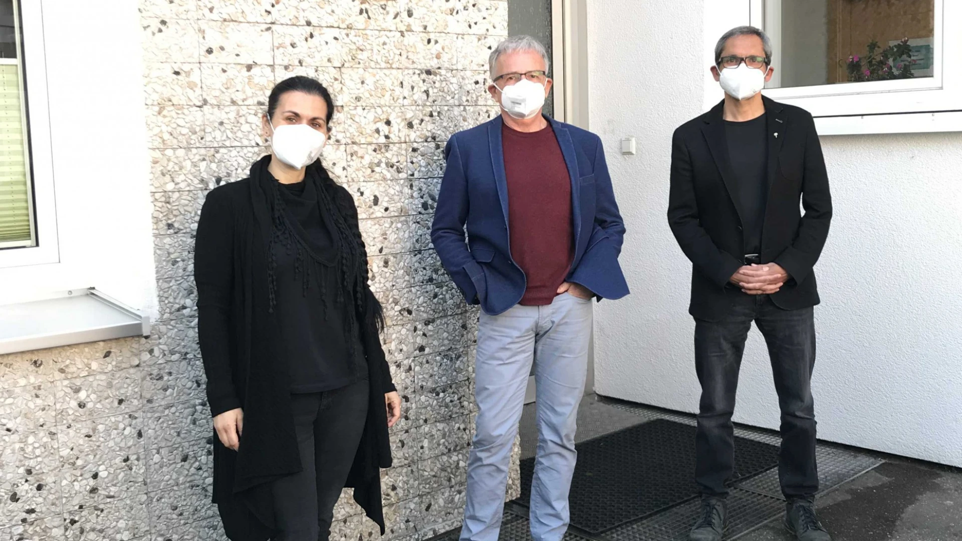 Drei Personen mit FFP3-Masken stehen vor einem Geb&auml;ude. Die Frau links tr&auml;gt schwarze Kleidung, der Mann in der Mitte ein dunkelrotes Oberteil mit blauem Sakko und heller Hose und der Mann rechts ist ebenfalls in schwarz gekleidet.
