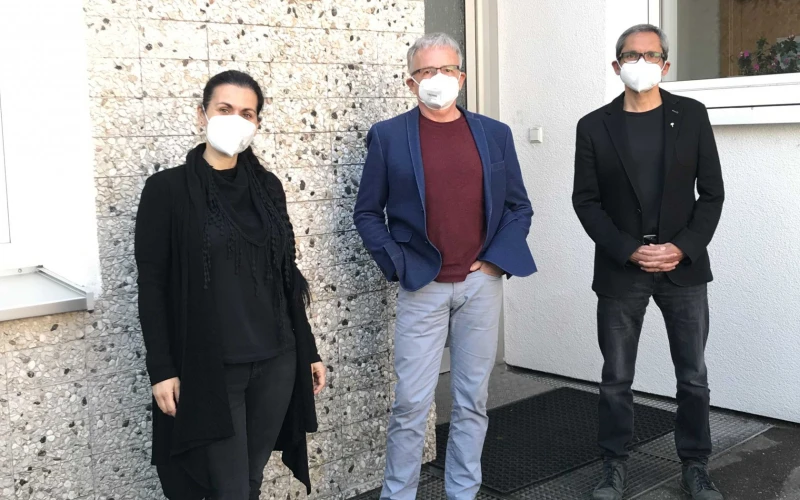 Drei Personen mit FFP3-Masken stehen vor einem Geb&auml;ude. Die Frau links tr&auml;gt schwarze Kleidung, der Mann in der Mitte ein dunkelrotes Oberteil mit blauem Sakko und heller Hose und der Mann rechts ist ebenfalls in schwarz gekleidet.