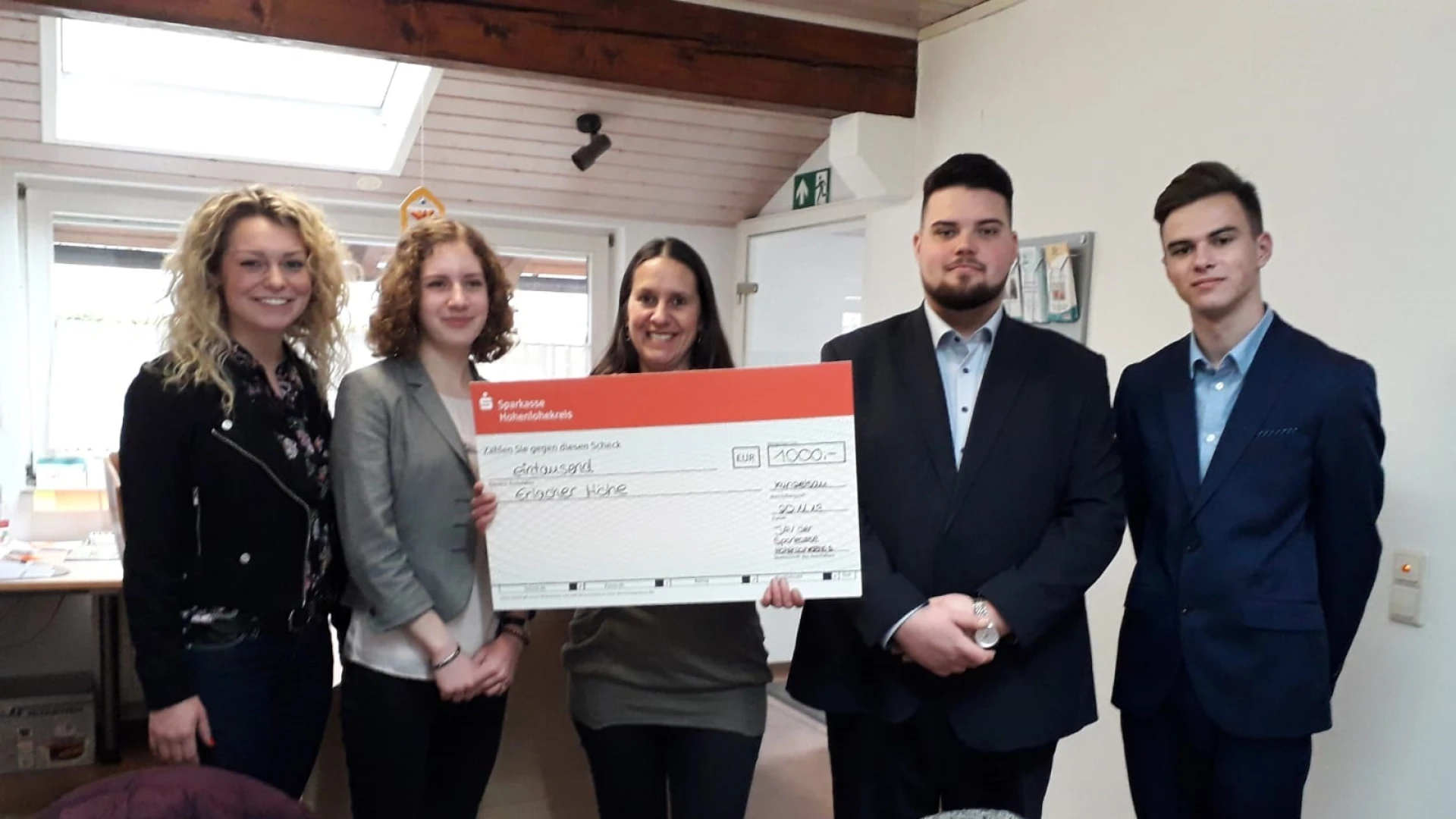 F&uuml;nf Personen befinden sich in einem Raum mit St&uuml;hlen, Tischen und Fenstern und schauen direkt in die Kamera. Die Frau in der Mitte h&auml;lt einen Scheck in der Hand.