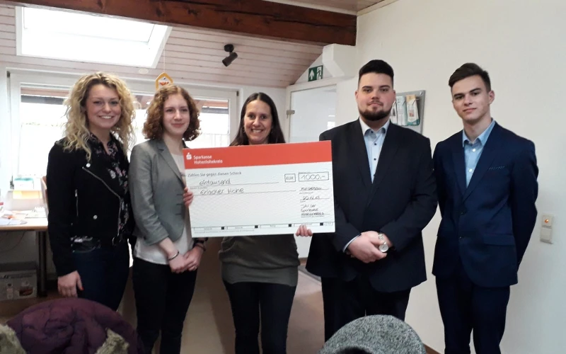 F&uuml;nf Personen befinden sich in einem Raum mit St&uuml;hlen, Tischen und Fenstern und schauen direkt in die Kamera. Die Frau in der Mitte h&auml;lt einen Scheck in der Hand.