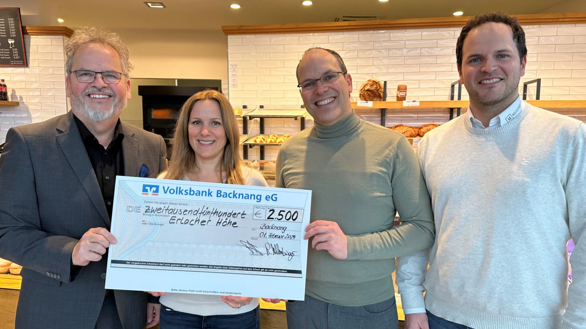 Vier Personen stehen in einem B&auml;ckerladen und l&auml;cheln in die Kamera. Im Hintergrund sind Regale mit verschiedenem Geb&auml;ck zu sehen ist. Ein Spendenscheck in H&ouml;he von 2500 Euro wird &uuml;bergeben.