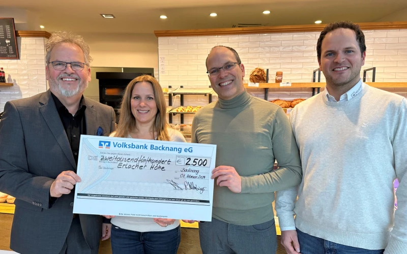 Vier Personen stehen in einem B&auml;ckerladen und l&auml;cheln in die Kamera. Im Hintergrund sind Regale mit verschiedenem Geb&auml;ck zu sehen ist. Ein Spendenscheck in H&ouml;he von 2500 Euro wird &uuml;bergeben.