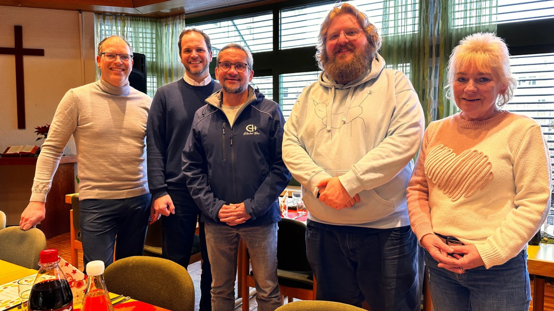 F&uuml;nf Personen stehen zusammen in einem Gemeinschaftsraum mit Holzdecke und gro&szlig;en Fenstern
