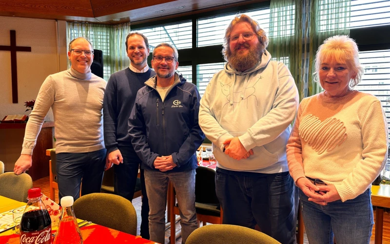 F&uuml;nf Personen stehen zusammen in einem Gemeinschaftsraum mit Holzdecke und gro&szlig;en Fenstern